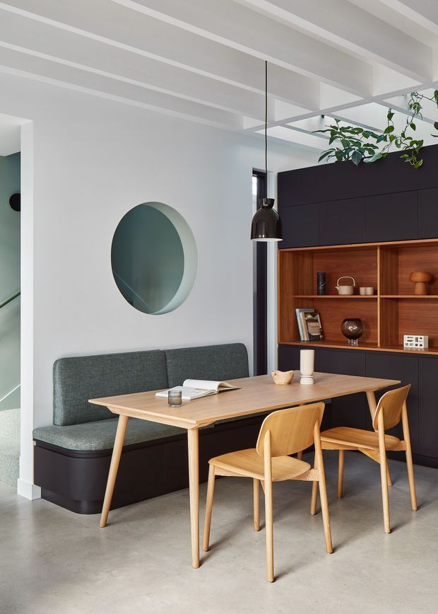 A refined dining nook with a built-in upholstered banquette, light timber dining table and matching chairs, and dark built-in cabinetry with warm timber open shelving. The room features a white ceiling with exposed linear beams, polished concrete flooring, a circular wall mirror, and a sculptural black pendant light. Indoor greenery adds a natural accent.