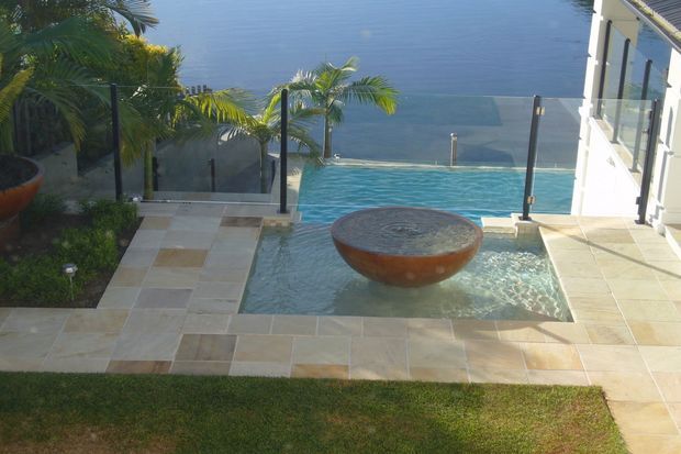 A luxury outdoor pool terrace overlooking a large body of water. The scene features a shallow tiled pool with an elevated circular stone or concrete water feature, transparent glass balustrades, pale stone paving, tropical palms, and landscaped planting. A modern white building edge is visible on the right.