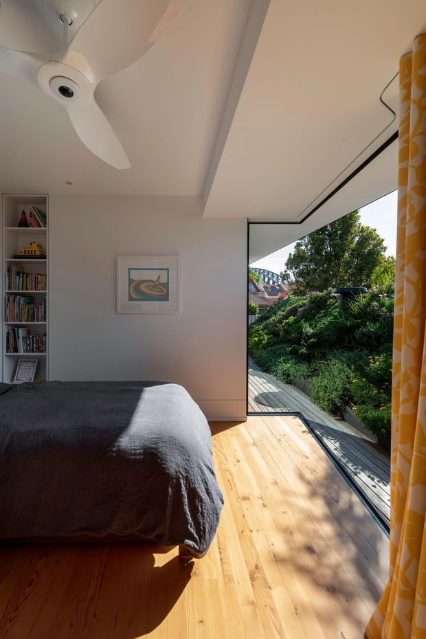 Bright, compact bedroom with a low platform bed, grey bedding, built-in open shelving, white walls and ceiling, and a large glazed opening leading to a narrow timber deck and leafy garden. A white ceiling fan, framed artwork, and bold yellow patterned curtains add character. Strong sunlight creates a warm, airy atmosphere.