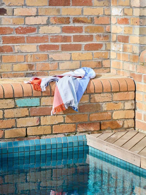Close-up view of a small swimming pool beside a rustic exposed-brick wall and brick bench, with a pale timber deck or pool edge at the right. The pool has vivid turquoise square tile lining and clear reflective water. A striped multicolour towel is draped over the brick bench, adding a casual outdoor character.