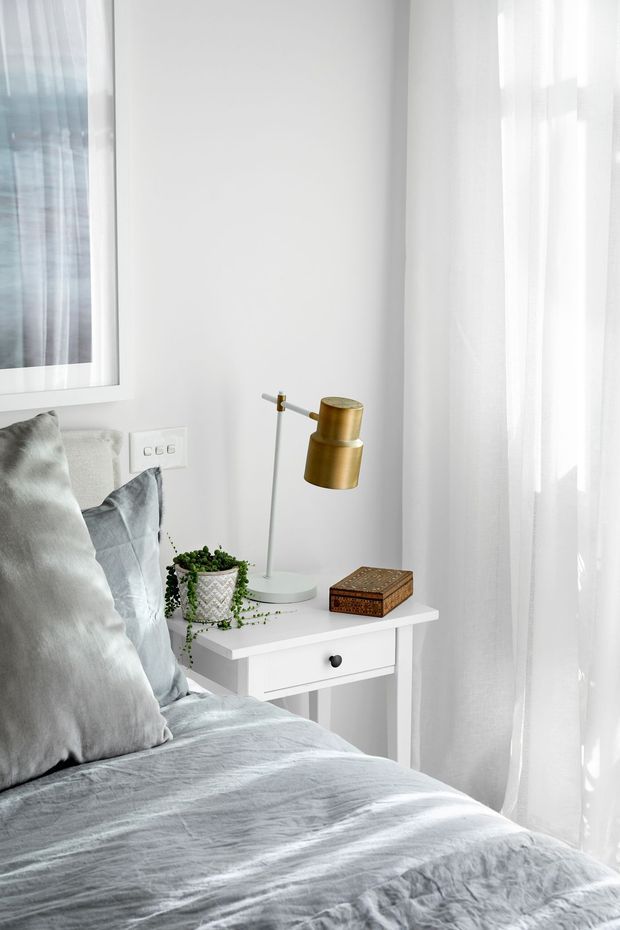 Bright, softly styled bedroom corner with a white bedside table, brass desk lamp, trailing potted plant, decorative wooden box, and layered grey bedding and pillows. Sheer white curtains diffuse abundant daylight beside the bed, creating a calm, minimalist atmosphere.