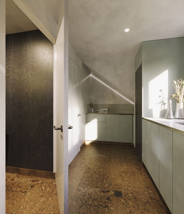 A narrow contemporary kitchen viewed through an open doorway, featuring muted green-grey flat-panel cabinetry, a dark tiled splashback, a long stone or terrazzo benchtop, and a warm brown textured floor. A sloped ceiling and angled recessed LED strip create a distinctive geometric form, while sunlight illuminates the side counter and decorative vases. An adjacent dark textured wall is visible at left.