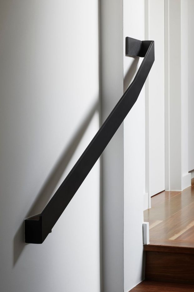 Close-up architectural detail of a matte black angular handrail mounted diagonally on a white wall beside a short flight of dark timber stairs. The image emphasizes the geometric metal rail, clean white wall surfaces, sharp shadows, and warm timber flooring and stair treads in the background.