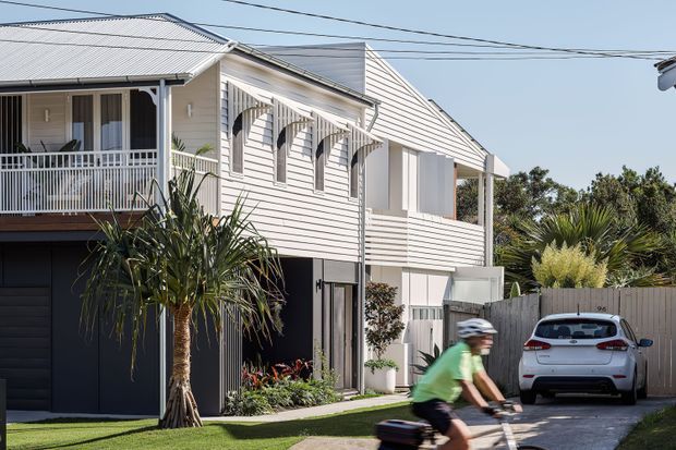 Exterior view of a narrow, multi-level coastal-style residence with pale horizontal weatherboard cladding, a steep gabled roof, covered upper balcony, decorative white window awnings, and a dark garage at ground level. A driveway, parked car, tropical planting, overhead utility lines, and a cyclist are visible in the foreground and surroundings.