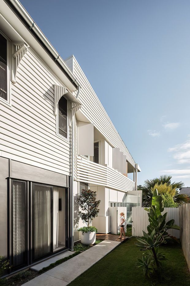 A bright exterior view of a narrow multi-storey residential building with white horizontal weatherboard cladding, projecting balconies and sun-shading panels, decorative window detailing, dark-framed glazed doors, and a landscaped private side courtyard with a paved walkway and tropical planting.