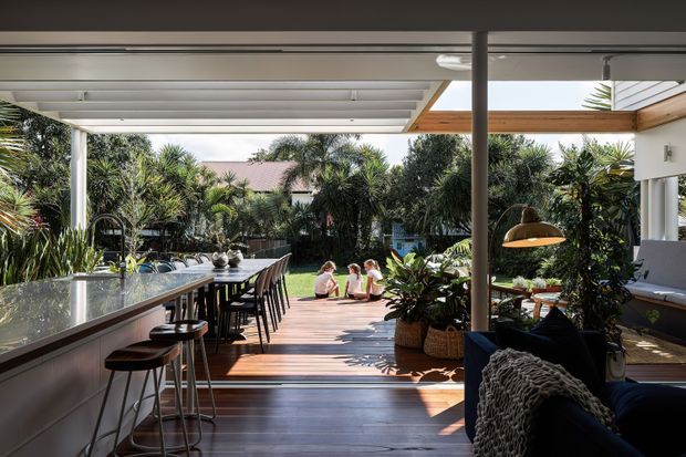 A spacious covered outdoor entertaining area connected to a contemporary home, featuring a long island-style outdoor kitchen or bar with stools on the left, a dining table and chairs, timber decking, lounge seating, and lush tropical landscaping surrounding a small pool or water feature. The pergola has a slatted roof and warm timber-lined sections, creating a bright indoor–outdoor living space.
