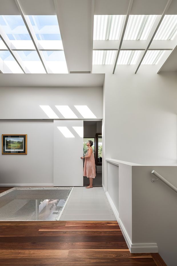 Bright contemporary interior hallway and landing with a stair opening in the foreground. The space features a dramatic high ceiling with extensive translucent skylight panels and exposed structural beams, white walls and joinery, dark timber flooring, pale tiled flooring, and a partially open sliding door. A framed artwork and a person provide scale, while strong daylight creates geometric patterns across the walls and floor.