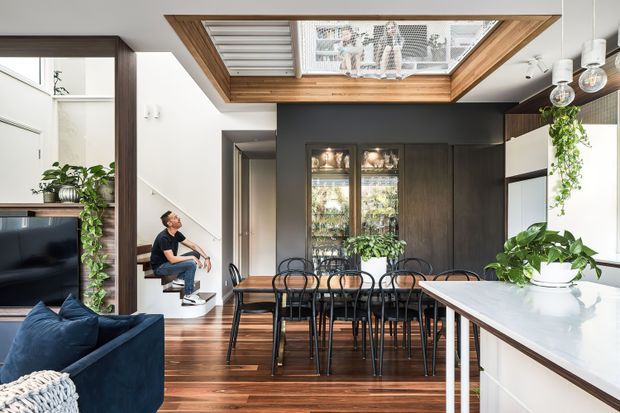 A bright, contemporary open-plan kitchen and dining area with a dramatic timber-framed skylight above the dining table. Dark timber cabinetry and a charcoal feature wall contrast with white walls and a pale kitchen island benchtop. The space includes a long dining table with black chairs, integrated shelving, indoor plants, timber flooring, and a partially visible navy sofa and television area. A seated person and a compact stair or reading nook appear at the side.