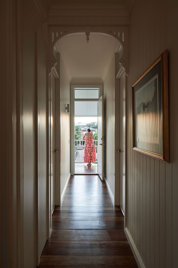 A narrow, warmly lit interior hallway with dark polished timber flooring, vertical timber wall panelling, white trims, and an ornate decorative arched opening. Several closed doors line the corridor, while an open glazed door at the far end leads to a small balcony or exterior landing. A framed artwork hangs on the right wall.