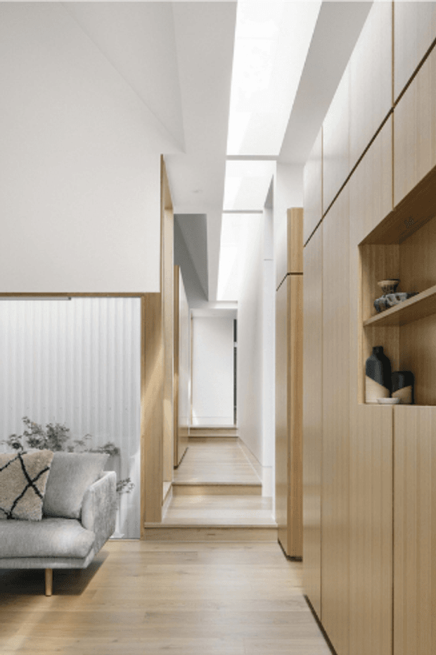 Bright, minimalist residential hallway with pale timber flooring and extensive integrated timber cabinetry. A short flight of steps leads to a raised corridor beneath a long skylight or clerestory, while a partial living area with a light grey sofa is visible at left. Built-in shelving displays a few decorative objects, and the overall palette is warm, light, and restrained.