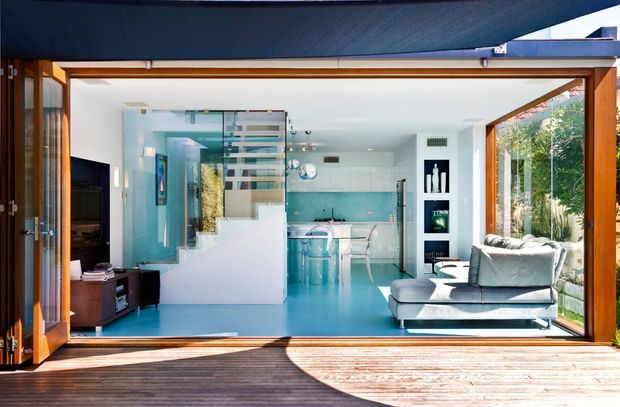 Bright, open-plan contemporary interior viewed from a timber deck through fully opened glazed timber doors. The room combines a living area with a sofa and media unit, a compact kitchen at the rear, and a glazed stair enclosure with floating stair treads. Strong turquoise-blue flooring and backsplash contrast with white walls, pale cabinetry, and warm timber framing. Large openings and extensive glazing connect the interior to the deck and garden.
