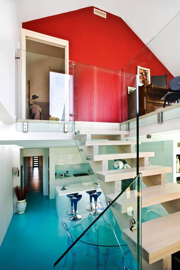 A bright, multi-level contemporary interior viewed from a stairway, featuring a vivid red upper wall, turquoise flooring, pale timber open-riser stairs and platforms, and clear glass balustrades. The lower level contains a compact white kitchen with a peninsula, cooktop, stools, and a transparent dining table with chairs. An upper-level landing and bedroom or dressing area are visible, with dark timber cabinetry and artwork.