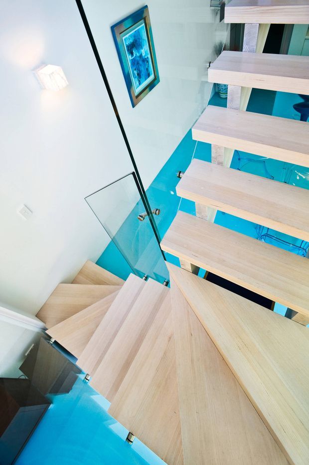 Interior view looking down through a contemporary open-riser stairway with pale timber treads, a glass balustrade, and bright turquoise flooring or surfaces below. White walls, a framed artwork, and visible structural details create a clean, modern architectural composition.