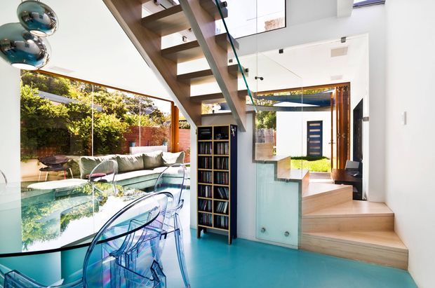 Bright contemporary open-plan interior featuring a dining area with a reflective glass table and transparent chairs, a turquoise polished floor, a floating timber-and-metal staircase, built-in shelving, and large glazed openings connecting to a landscaped courtyard and entry. White walls, skylights, and abundant natural light create an airy, modern atmosphere.