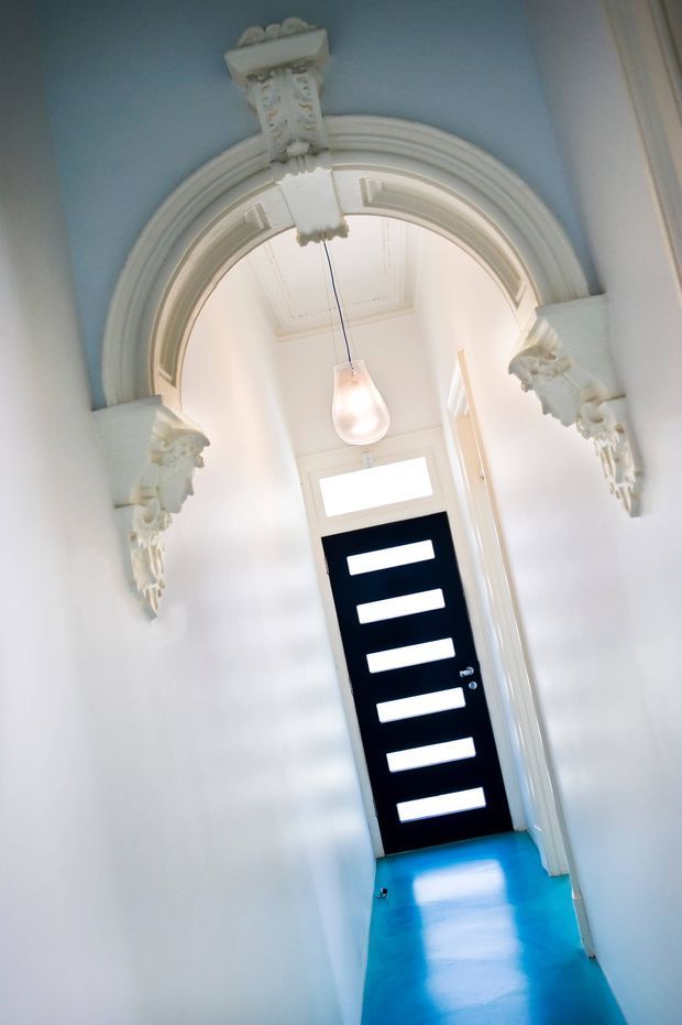 A narrow residential hallway viewed toward a striking black front door with six horizontal glazed panels and a transom window. The corridor features highly ornate white plaster architraves, decorative corbels and classical detailing, pale blue upper wall sections, white walls and trim, a glossy vivid blue floor, and a single pendant light.