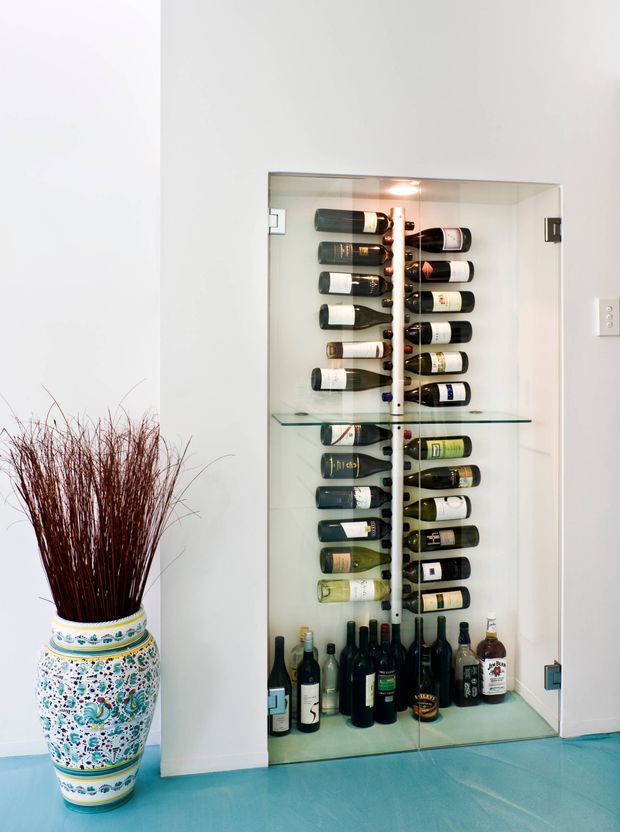A tall built-in glass-front wine display and storage cabinet occupies a recessed niche in a white wall. Numerous wine bottles are stored horizontally on internal racks, with larger bottles standing on the bottom. The cabinet has clear glass shelving and doors with dark metal hinges and hardware. A large patterned ceramic floor vase with tall dried branches sits beside it, and the surrounding floor is turquoise-blue. The space is bright, clean, and primarily focused on wine storage.