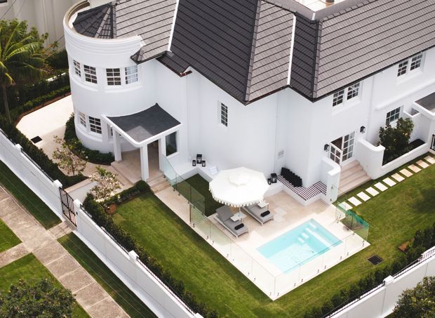 Aerial view of a large white multi-storey residence with a steep dark grey tiled roof, curved bay tower, white-framed windows, manicured lawns, paved outdoor terrace, and a small rectangular swimming pool within a fenced property.