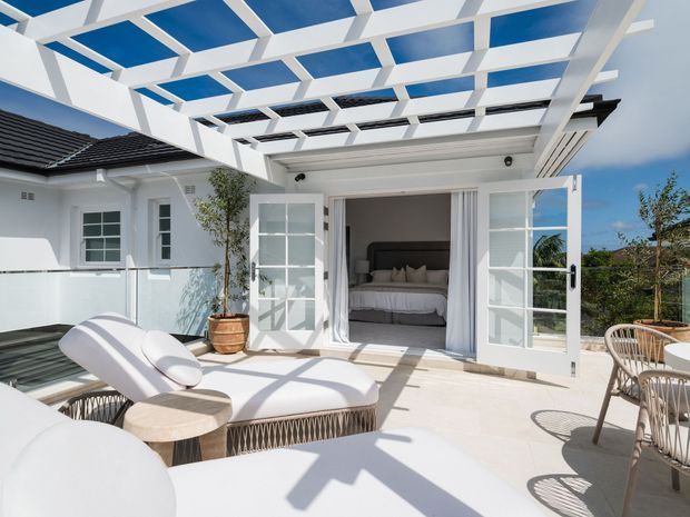 Bright white outdoor terrace adjoining a bedroom, with a modern pergola casting geometric shadows, folding glass-panel doors, pale tiled paving, sun loungers, and a small outdoor dining setting. The open doors reveal a calm, neutral bedroom interior.