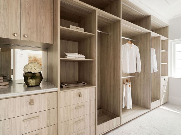 A bright, built-in walk-in wardrobe with full-height light timber cabinetry, open hanging sections, shelves, drawers and upper cupboards. A small stone benchtop and decorative display niche appear at left, while white clothing and folded items are stored throughout. The space has a pale carpeted floor, white walls and strong natural light from a window at right.