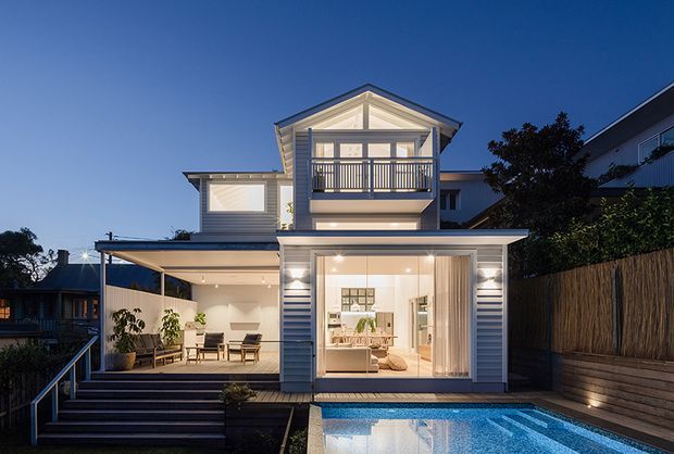Twilight exterior view of a contemporary multi-level coastal-style home with light weatherboard cladding, a prominent upper gabled balcony, covered outdoor living deck, and large glazed openings. A swimming pool occupies the foreground, with warm interior lighting visible through the ground-floor glazing.