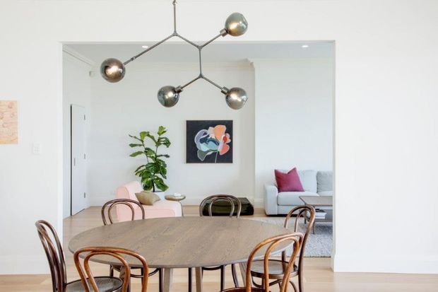 Bright open-plan dining and living area with a large oval wood dining table and curved bentwood chairs in the foreground. A sculptural multi-arm pendant light hangs above the table, while the adjoining living area contains a pale sofa, pink accent cushion, plant, and abstract artwork. The space has white walls, light timber flooring, simple white door and trim, and a large wall opening creating visual connection between the rooms.