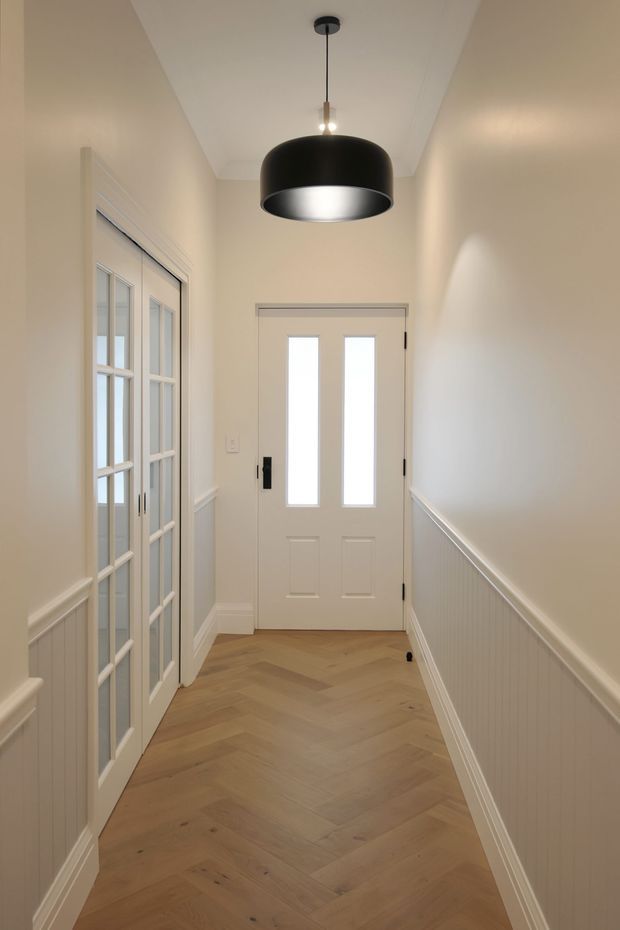 A narrow, bright residential entry hall with warm off-white walls, white vertical wall panelling and trim, a white glazed front door, and matching internal French doors. The floor is laid in a natural light timber herringbone pattern. A large black pendant light provides a contemporary focal point against the simple ceiling.
