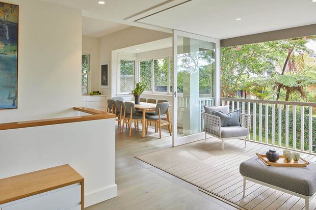 Bright, open-plan contemporary interior connecting a dining area and kitchen with a covered timber deck through large glazed sliding doors. The space features white walls and cabinetry, light timber flooring and joinery, a timber dining table with upholstered chairs, and a woven outdoor-style lounge chair and ottoman overlooking leafy vegetation. Recessed ceiling lighting and a slim linear ceiling vent add to the clean modern fit-out.