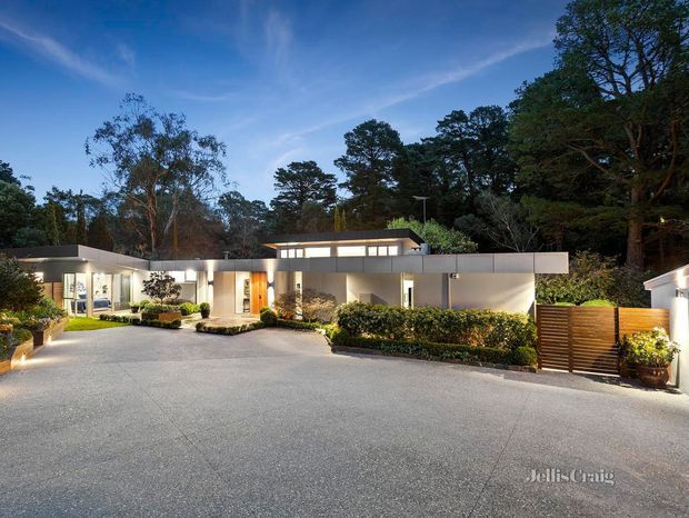 Wide dusk exterior view of a low, flat-roofed modern residence set within a landscaped, tree-filled site. The home has white rendered walls, extensive glazing, warm interior lighting, a broad paved driveway or forecourt, manicured hedges, planters, and a timber slatted gate.