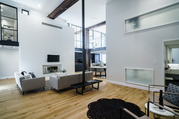 Bright, open-plan contemporary living room with pale timber flooring, white walls, exposed dark timber beams, black-framed glazing, and a double-height industrial-style void. The room contains a grey sofa and armchairs arranged around a low black coffee table, with built-in shelving and a wall-mounted television. A dining area and kitchen are visible beyond the living space, while a bathroom is visible through the doorway at right.