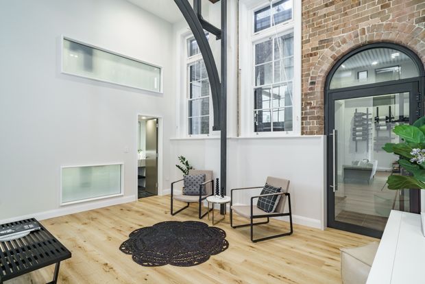 Bright, double-height contemporary interior with exposed dark steel structural framing, tall white-framed windows, exposed red-brown brick walls, and pale timber flooring. A glass-panelled arched doorway opens to an adjacent space, while two modern lounge chairs, a small side table, patterned rug, and indoor plants create a compact seating area.