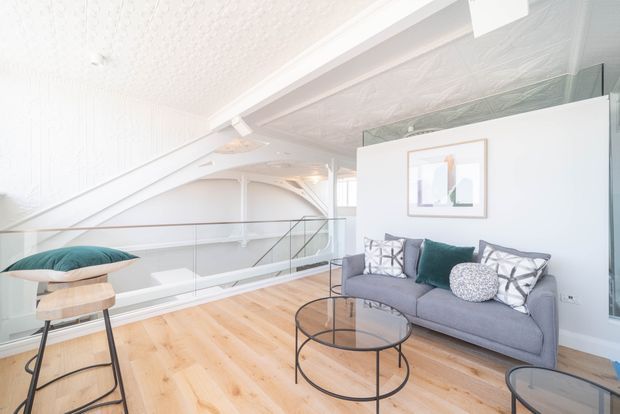 Bright, airy loft-style living area with a grey sofa, teal and patterned cushions, round glass coffee tables, and light timber flooring. The room features a sloping textured ceiling, exposed white structural beams, glass balustrading around a stair or landing void, and large glazing, creating an open mezzanine-like setting.