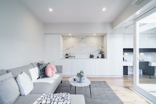 Bright, contemporary open-plan living area with a pale grey sectional sofa, layered cushions, a round coffee table, and a textured grey rug. White built-in cabinetry and a dramatic white veined stone feature backsplash form a compact kitchen or bar area at the rear. Light timber flooring, recessed ceiling lights, and large glazed doors create an airy connection to an adjacent dining or kitchen space.