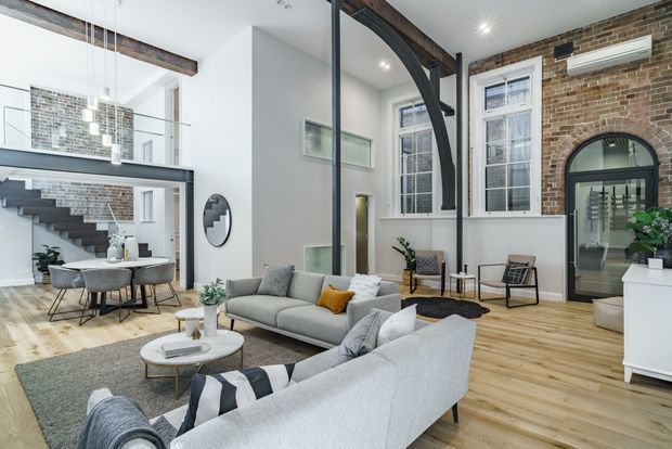 A bright, open-plan loft-style living room with a large grey sectional sofa, coffee tables, dining area, and double-height volume. Exposed dark timber beams, black-framed internal glazing, a dramatic curved black-framed partition, tall multi-pane windows, exposed brick walls, and a mezzanine staircase create a refined industrial character. Light timber flooring and white walls balance the darker structural elements.