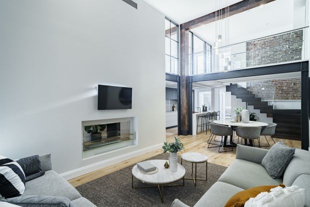 Bright, spacious contemporary loft-style living room with a high ceiling and double-height glazing. Two light grey sofas face a wall-mounted television and recessed fireplace, with a round marble coffee table and textured rug in the foreground. The open-plan dining area, exposed dark structural beams, black-framed glazing, brick feature wall, and black staircase leading to a mezzanine are visible beyond.
