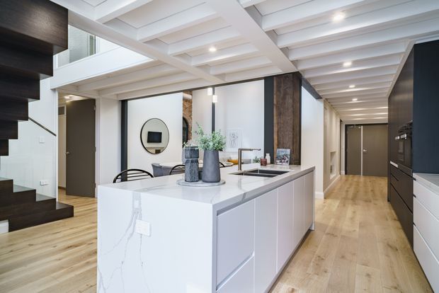 A bright contemporary open-plan kitchen and hallway with a large white waterfall island, integrated dark cabinetry, pale timber flooring, and a white coffered ceiling. Dark timber stair treads and a substantial timber-clad structural column add contrast, while a dining area and double-height void are visible beyond the island.