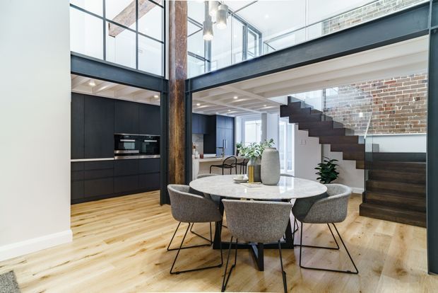 A bright, contemporary double-height dining and kitchen interior in a converted industrial-style space. A round marble dining table with upholstered chairs sits on pale timber flooring, adjacent to a matte black kitchen with integrated appliances. Exposed brickwork, dark steel structural beams, a substantial timber post, and a floating dark timber staircase contribute strong industrial character. Upper-level glazing and a mezzanine-like void enhance the airy vertical scale.