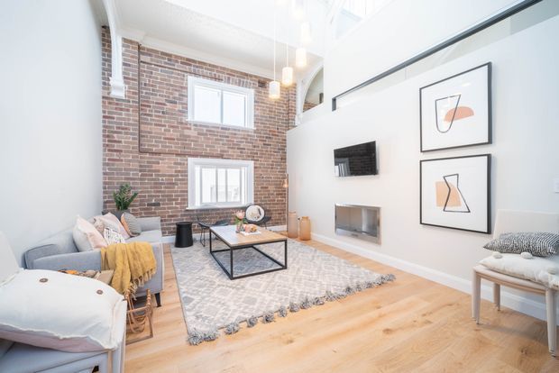 Bright, airy contemporary living room with a double-height ceiling and mezzanine-style upper opening. The room features an exposed red-brick wall with two windows, white walls, pale timber flooring, a grey sofa, patterned rug, black metal coffee table, wall-mounted television and framed abstract artwork. Pendant lights hang in the tall space, and a partial white-painted brick arch is visible above the living area.