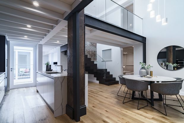 A bright, open-plan contemporary interior combining a kitchen, dining area and stairway beneath a double-height void. The space features white cabinetry and walls, a pale timber floor, a veined stone kitchen island, exposed dark structural beams, a black staircase, glass balustrading and a round wall mirror. A mezzanine level overlooks the dining area, with pendant lights providing focused illumination.
