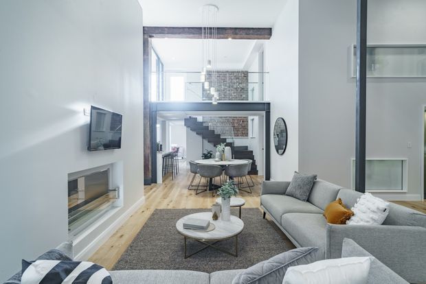 A bright, open-plan contemporary living room viewed toward a dining area and kitchen, with a large grey sectional sofa, round coffee table, textured rug, wall-mounted television, and built-in linear fireplace. Exposed dark timber beams, brickwork, black-framed glazing, and a sculptural open staircase create an industrial loft-like character. A dining table and kitchen are visible beyond the living area.