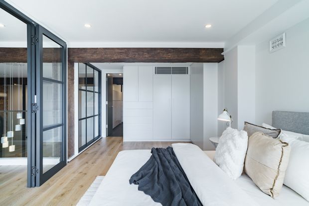 Bright contemporary bedroom with a large bed, layered neutral bedding, white built-in cabinetry, pale timber flooring, black-framed glazed partitions, and a prominent dark exposed timber beam across the ceiling. The room opens toward a glazed area and hallway, with recessed ceiling lighting and a small bedside lamp.
