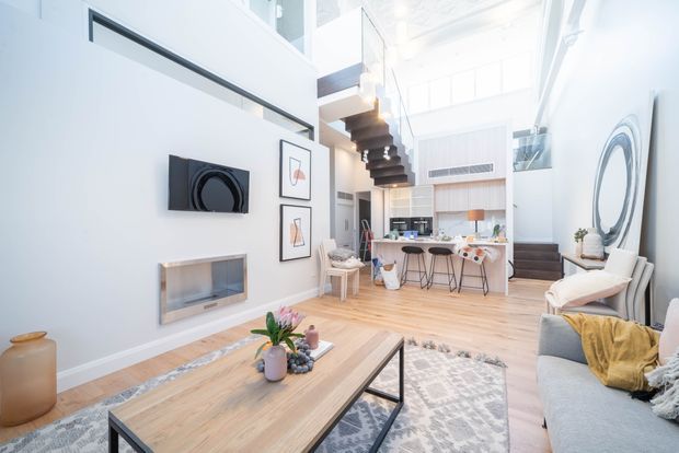 Bright contemporary open-plan living space with a double-height ceiling and mezzanine level. The room combines a light timber floor, white walls, a high-volume glazed upper level, a floating sculptural staircase, and an open kitchen with island seating. A large light-wood coffee table, neutral sectional sofa, patterned rug, framed artwork, and oversized circular wall mirror furnish the living area.