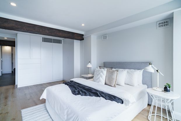 Bright, calm contemporary bedroom with a large upholstered bed, white bedding, pale grey walls, and built-in white wardrobe cabinetry. Light timber flooring, a dark timber-look ceiling beam, bedside tables, and layered task lighting create a clean, refined interior. A hallway and built-in storage are visible in the background.