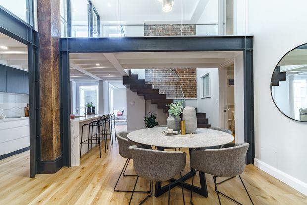 Bright contemporary open-plan dining area with a round stone-look dining table and upholstered chairs, positioned beneath a steel-framed mezzanine or upper-level opening. The space features exposed red brick, a dark timber-and-steel stair, warm timber flooring, large windows, and a visible kitchen area to the left. A round wall mirror reflects the stair and glazing.