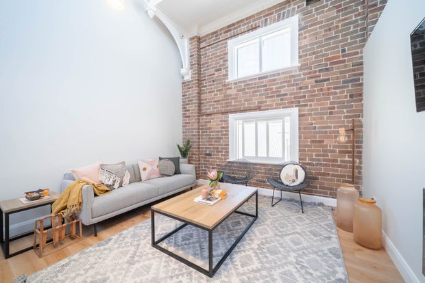 Bright living room with a pale grey sofa, patterned cushions, a large light patterned rug, and a rectangular timber coffee table with a black metal frame. The room features an exposed red brick feature wall with two white-trimmed windows, white plaster walls and ornate white ceiling cornices, plus minimalist black-framed occasional furniture and decorative vessels.