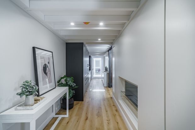 A bright, narrow residential hallway with warm timber flooring, white walls, and a white exposed-beam ceiling. A slim console with plants and artwork sits to the left, while a dark built-in joinery wall and a recessed fireplace or display opening line the corridor. Glazing and a door at the far end bring in natural light.
