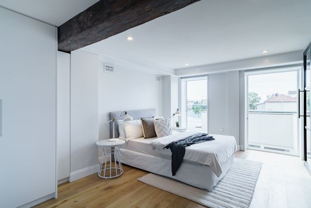 Bright contemporary bedroom with a low upholstered bed, layered neutral bedding, slim bedside tables, and a large textured rug on warm timber flooring. White walls and full-height glazing create an airy feel, while a dark exposed timber beam adds architectural contrast.