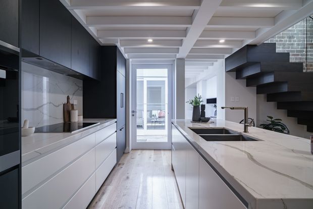 A refined contemporary galley kitchen with white and black cabinetry, a large white marble-look island, integrated appliances, and a dark floating staircase rising along the right wall. The room features a striking white coffered ceiling with recessed lighting, pale timber flooring, and a glazed door at the rear opening to a bright adjacent space.