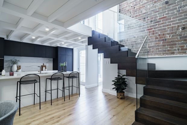 A bright, open-plan contemporary kitchen and stair hall. The kitchen features matte black full-height cabinetry, a white marble-look splashback and island bench with slim black metal bar stools. Pale timber flooring contrasts with a dark timber staircase, clear glass balustrade, exposed red brick wall, and a white coffered ceiling. A large skylight or clerestory opening brings in abundant natural light.