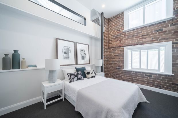 Bright contemporary bedroom with a double bed, white bedding, bedside tables and lamps, framed artwork, and built-in shelving. One wall features exposed red brick with large white-framed windows, while the opposite wall is painted white with a high internal clerestory-style opening. The room has dark carpet flooring and a clean, modern loft-like character.