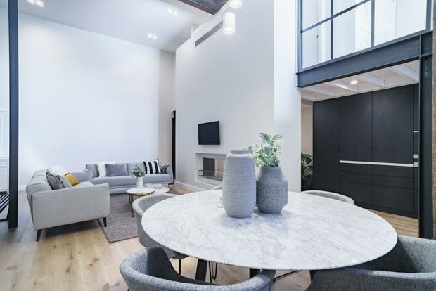 Bright contemporary open-plan living and dining area with a double-height ceiling and upper-level glazing. A round marble dining table with upholstered chairs dominates the foreground, while a light grey sofa arrangement, wall-mounted television, minimalist fireplace, indoor plants, and dark built-in cabinetry occupy the background. Pale timber flooring, white walls, black-framed glazing, and a restrained neutral palette create a refined modern interior.